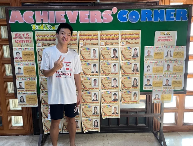 Anderson Tsai 站在 CPILS 校內 Achievers' Corner 公布欄前比 YA,欄上張貼 IELTS 與 TOEIC 達標學生名單